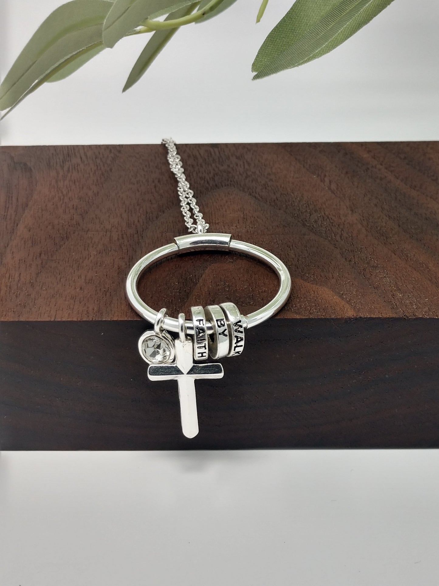 Close up of antique silver tone necklace with three charms one reads walk, one reads by and one reads faith. Small crystal charm and silver tone cross charm. Round silver tone ring on end of silver tone necklace
